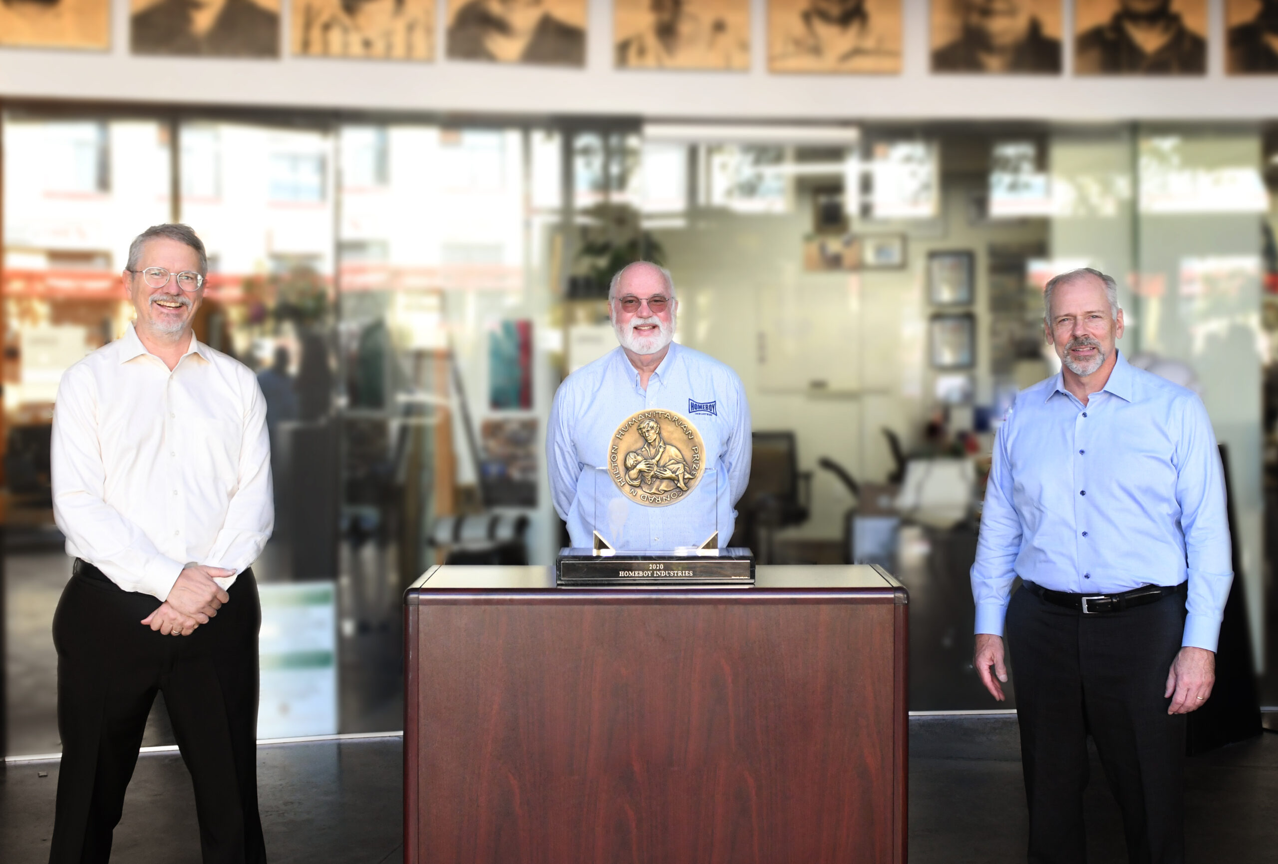 Three smiling men stand apart from each other in front of a glass building and behind an award.